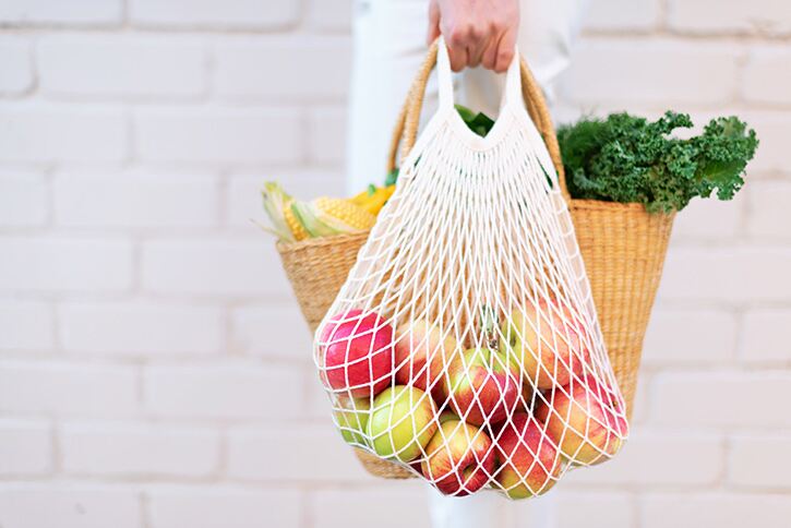 A hand holds a string bag of apples and a straw bag of kale and corn